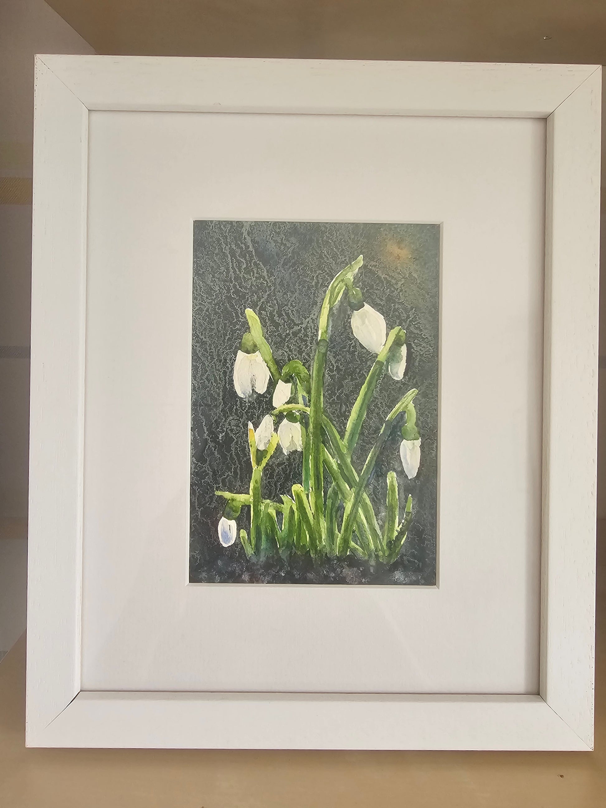 A framed art print depicting snowdrops with white blossoms and green stems against a dark textured background, presented in a white frame with a white mat.